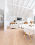 Open-plan lounge and dining room with vaulted white ceiling and natural decor elements, featuring a neutral modular sofa, round white dining table with upholstered chairs, dried floral arrangements, and wooden accents.