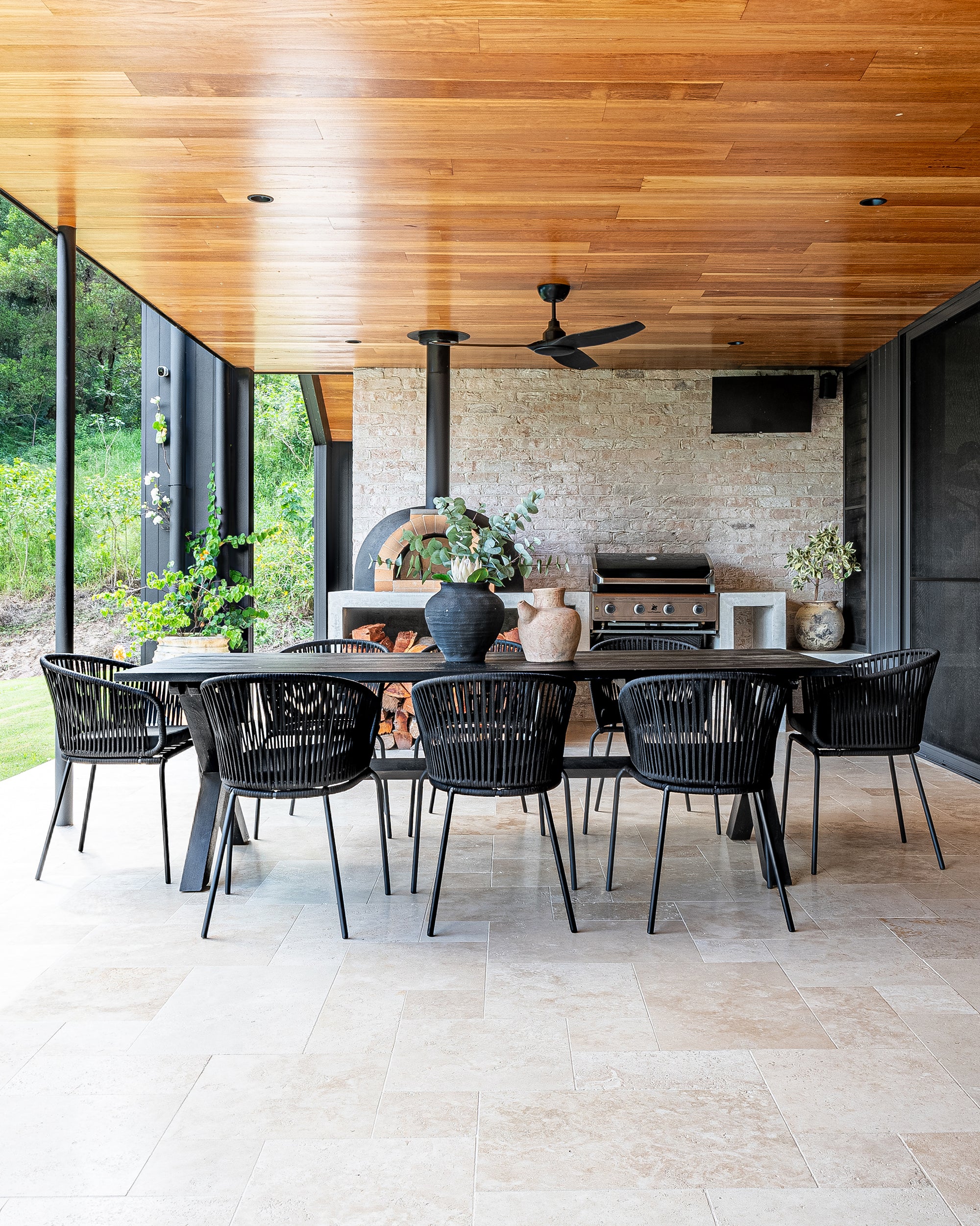 A stylish outdoor dining space featuring a black gable house with timber ceiling details, an open-plan living area, and a built-in wood-fired pizza oven. Surrounded by lush greenery, this modern retreat blends contemporary architecture with natural elements