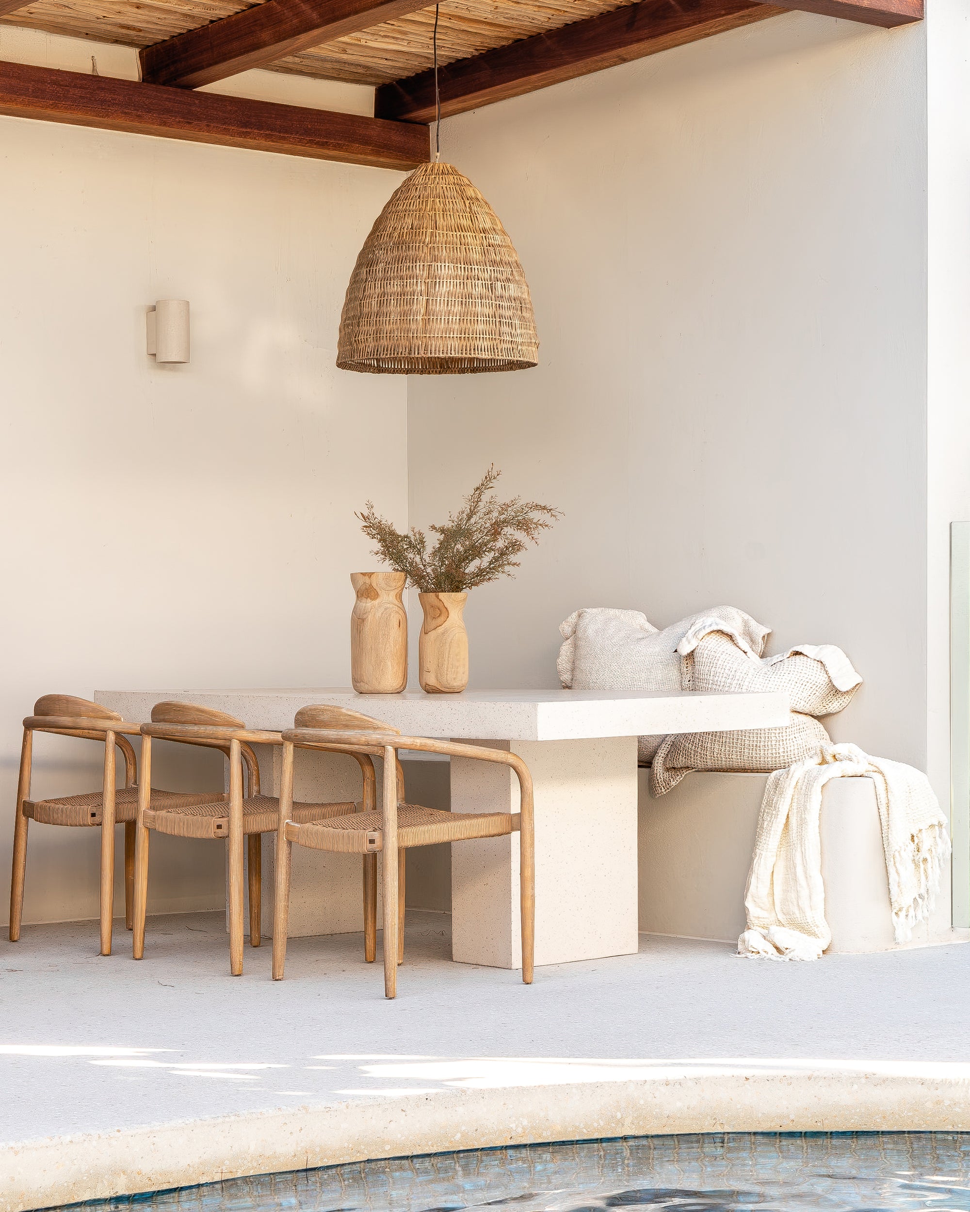 Outdoor dining setting featuring a teak chairs and travertine-look dining table beside a pool under a woven pendant light