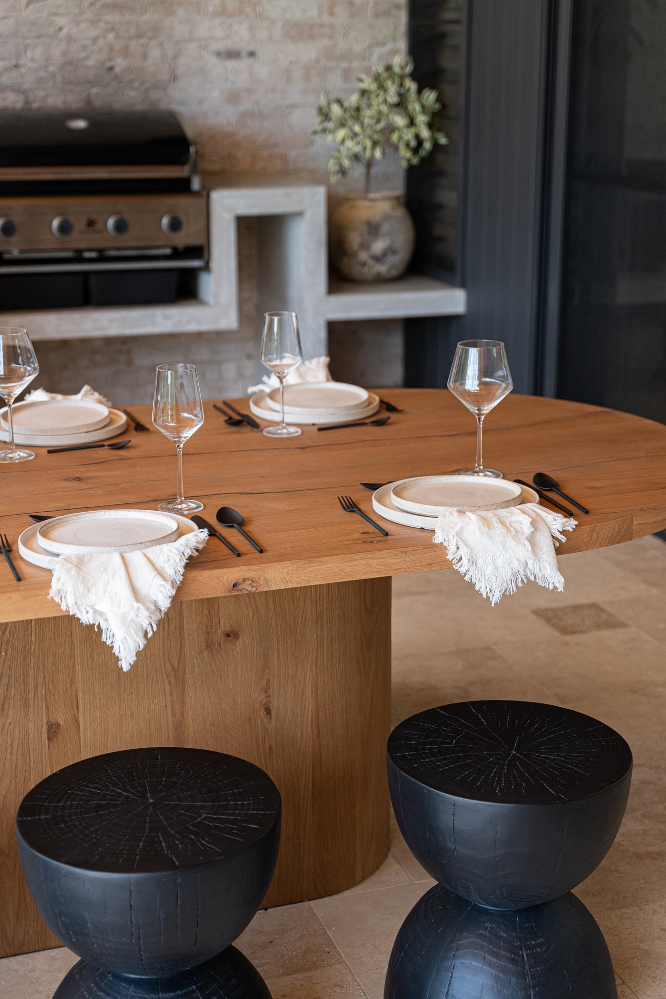 An elegant outdoor dining setup featuring a natural timber table with ceramic plates, matte black cutlery, fringed linen napkins, and sculptural blackwood stools, set against a rustic brick wall with a built-in barbecue