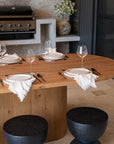 An elegant outdoor dining setup featuring a natural timber table with ceramic plates, matte black cutlery, fringed linen napkins, and sculptural blackwood stools, set against a rustic brick wall with a built-in barbecue