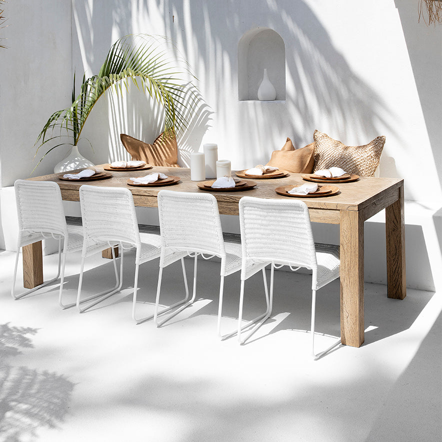 A serene outdoor dining setup by Kulala Living featuring a solid wood dining table paired with white wicker chairs, styled with earthy-toned cushions, ceramic vases, and natural tableware under soft dappled sunlight