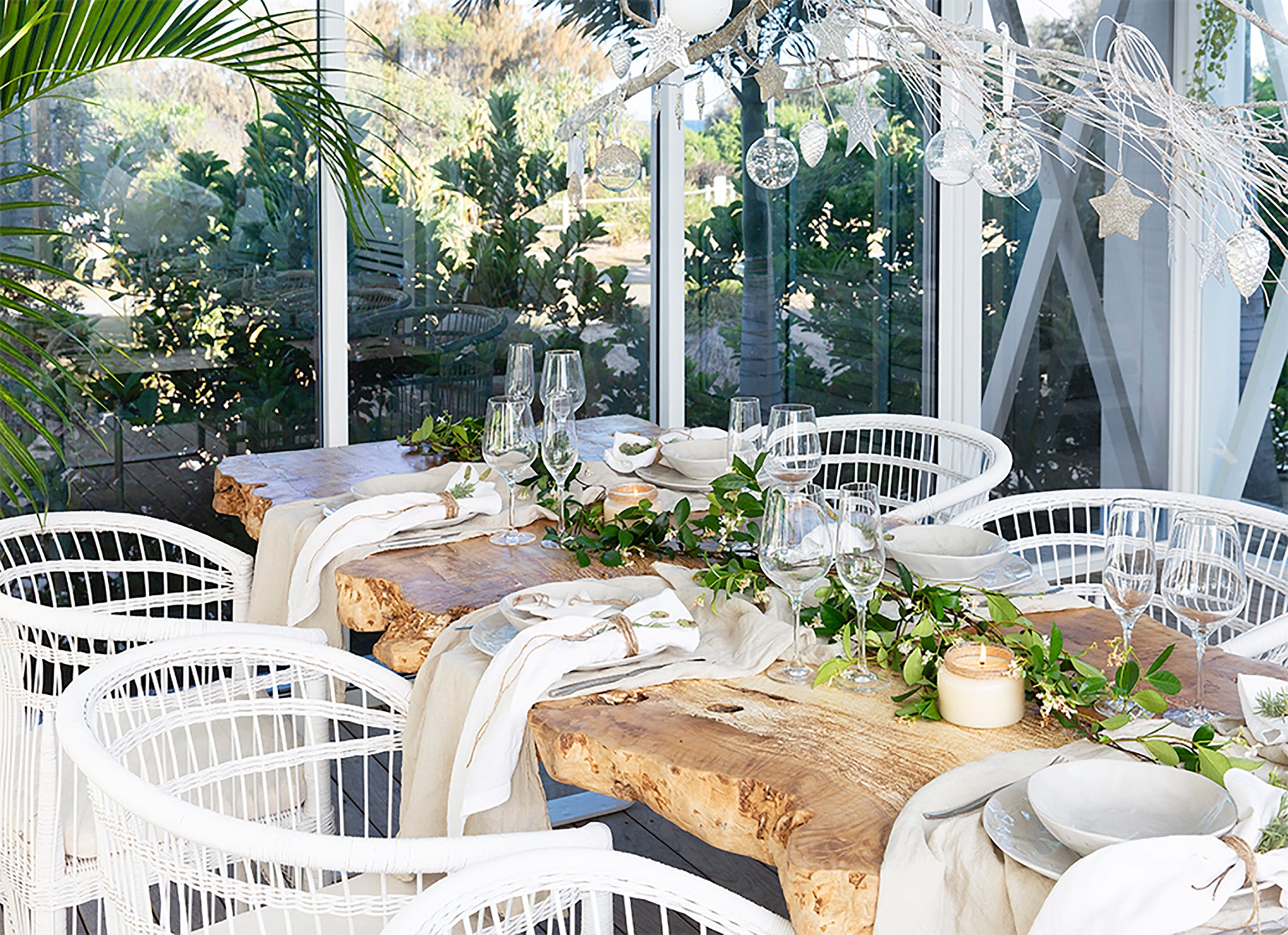 A beautifully styled outdoor dining space featuring a natural live-edge wooden table, white woven chairs, and elegant table settings with greenery and glassware, creating a refined yet relaxed entertaining area