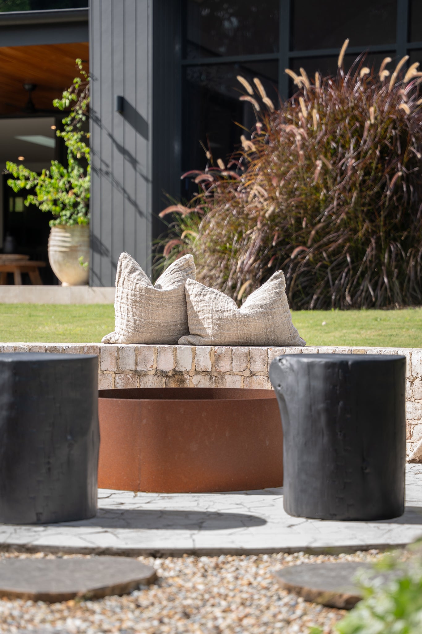 A modern outdoor setting featuring a rusted fire pit surrounded by black timber stools and textured linen cushions on a rustic brick ledge. The natural tones and organic textures blend seamlessly with the lush greenery, creating a serene and inviting space for outdoor relaxation