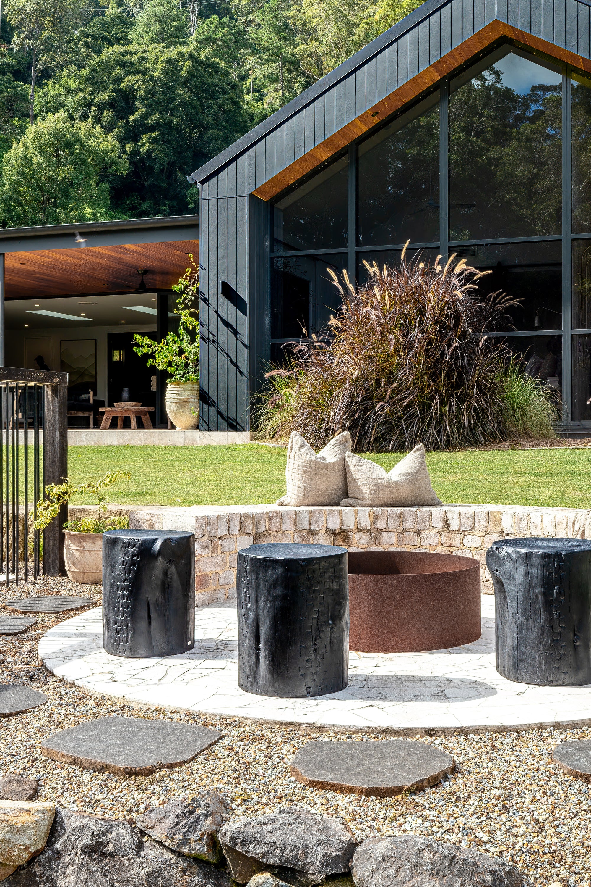A serene outdoor setting featuring KULALA Living’s black wooden stools arranged around a rusted steel firepit. Set against a modern black-clad home with lush greenery, this space blends contemporary design with natural elements for a stylish retreat. Neutral linen cushions add comfort to the inviting ambiance