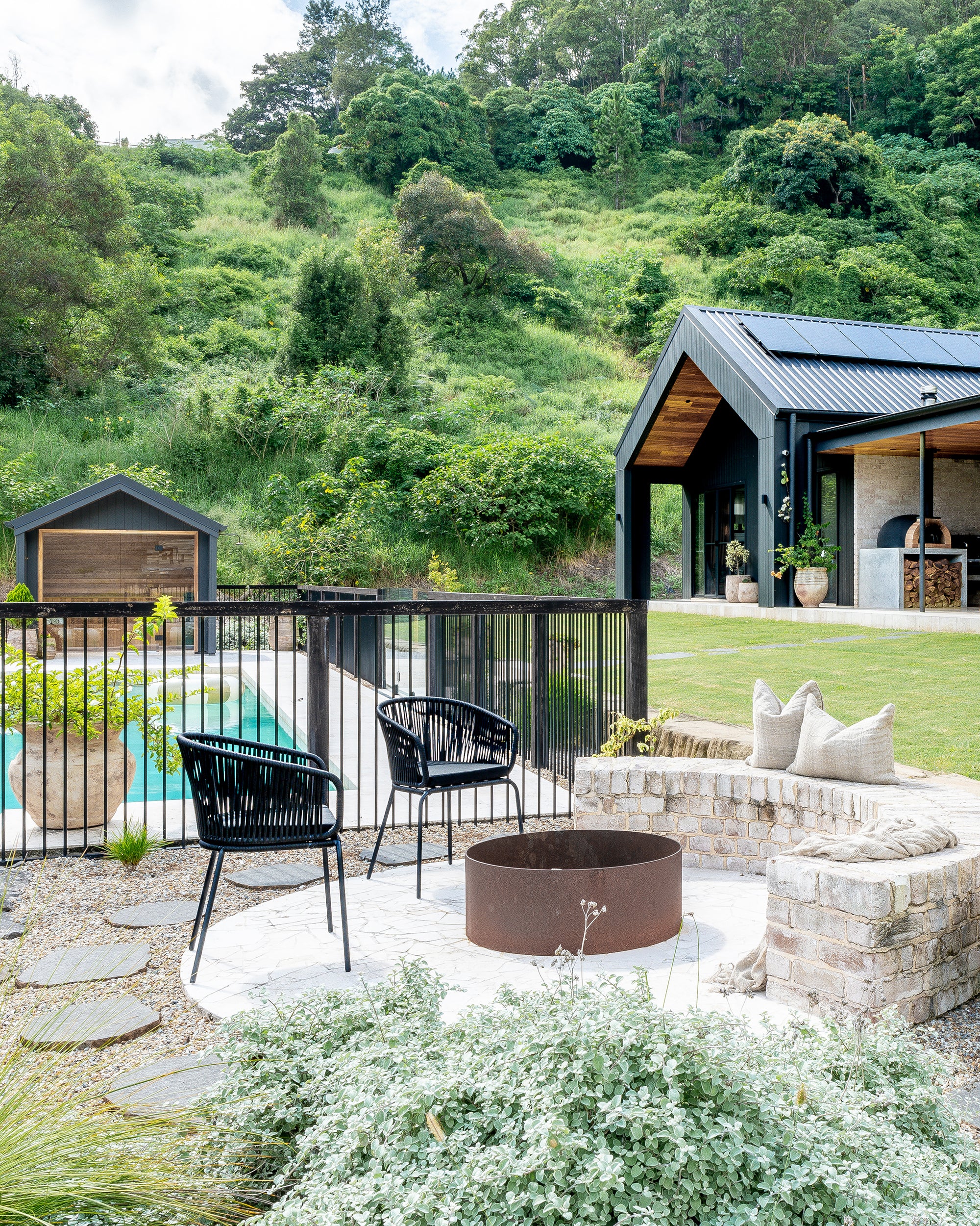 A tranquil outdoor firepit setting featuring modern black woven chairs, a rustic brick bench with linen cushions, and a lush hillside backdrop. This serene poolside space complements the contemporary black-clad home and open outdoor kitchen, creating a stylish and inviting retreat