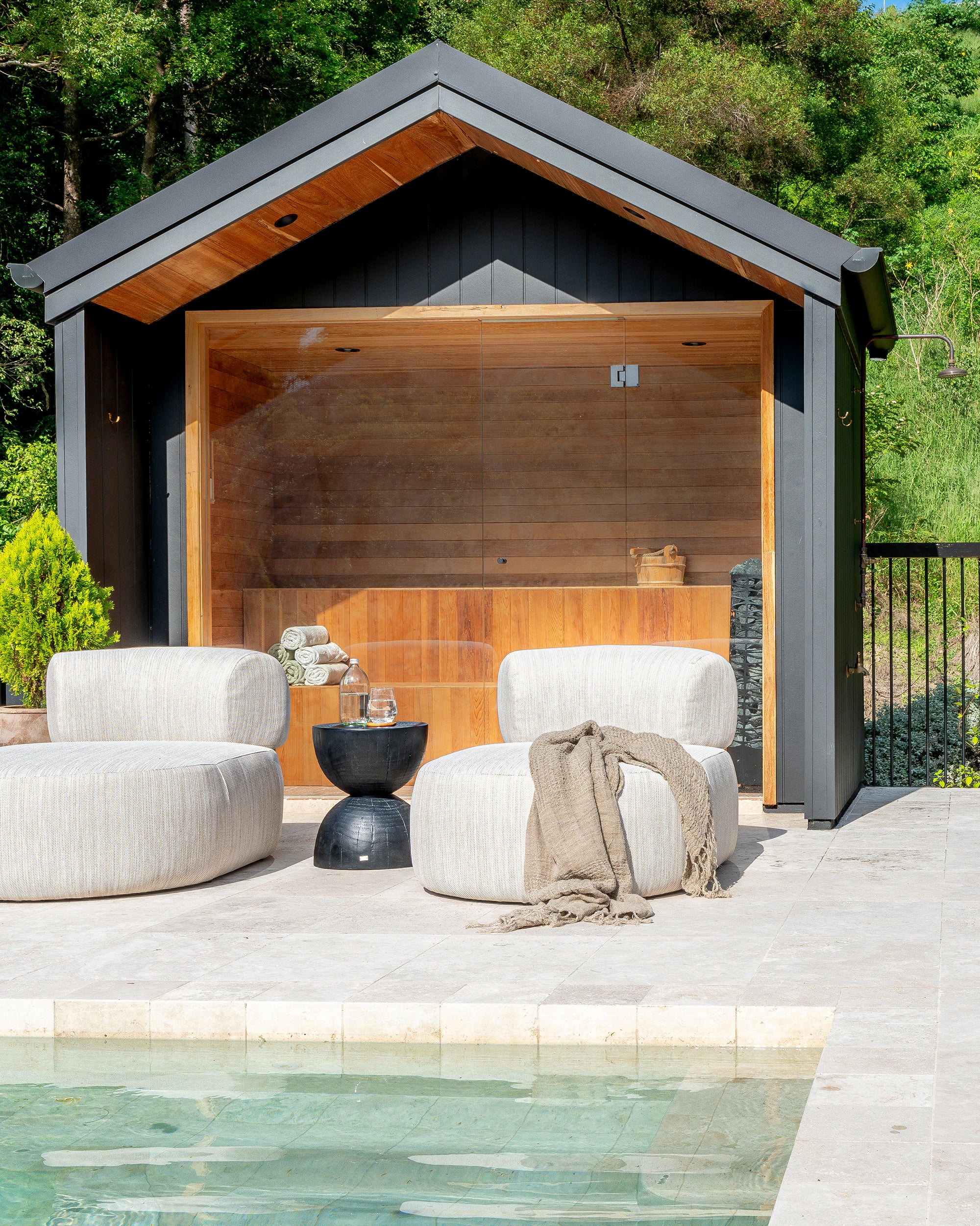Outdoor setting with two rounded neutral loumgers and a black hourglass side table in front of a timber-lined sauna, positioned beside a tranquil pool area.