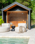 Outdoor setting with two rounded neutral loumgers and a black hourglass side table in front of a timber-lined sauna, positioned beside a tranquil pool area.