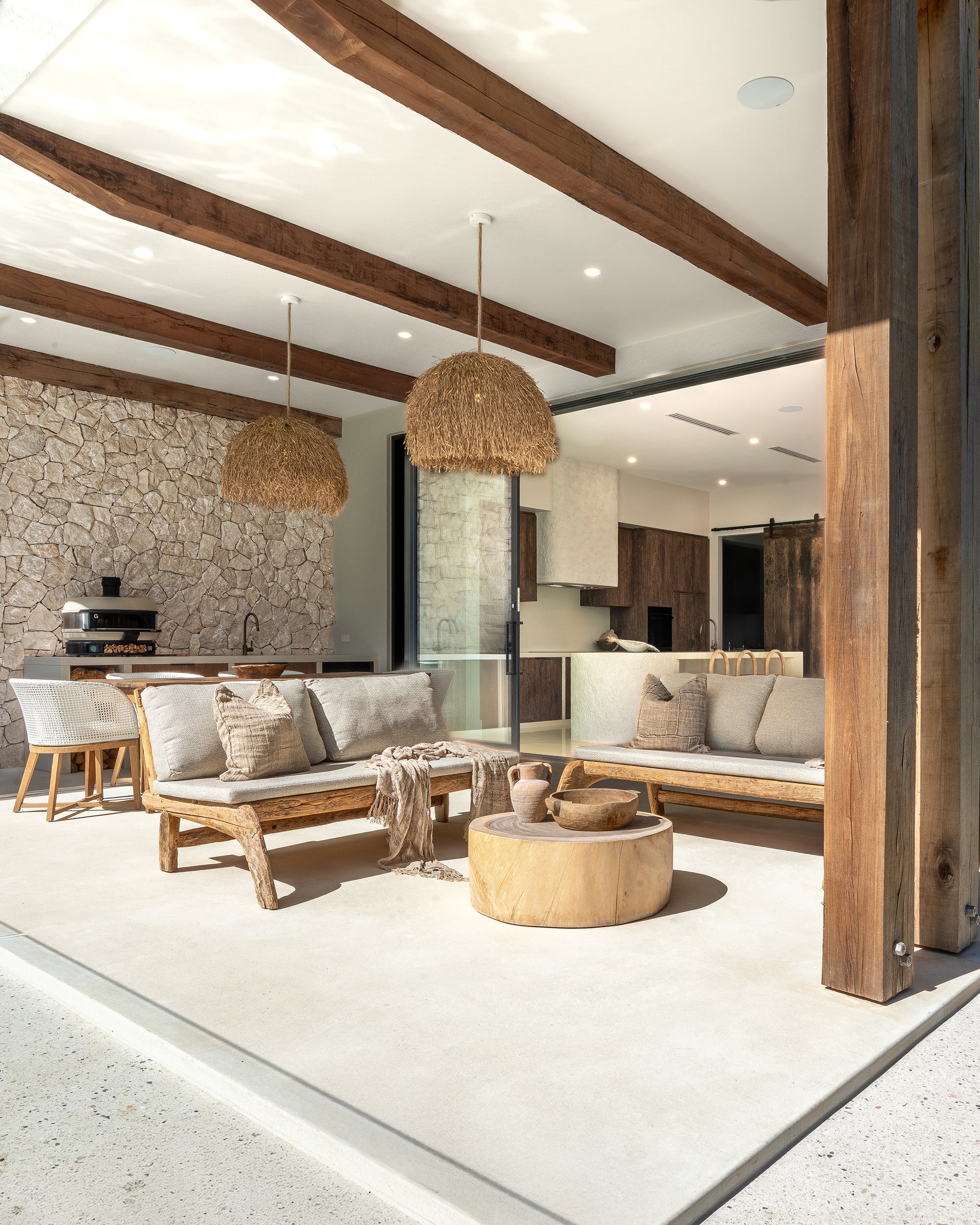 Ooutdoor living area with teak sofas, timber coffee table, stone feature wall, and natural cushions and throw beside an open kitchen.
