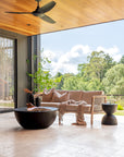 Covered outdoor patio featuring a teak-framed sofa with textured cushions, black round coffee table, and garden views framed by a timber ceiling and ceiling fan.