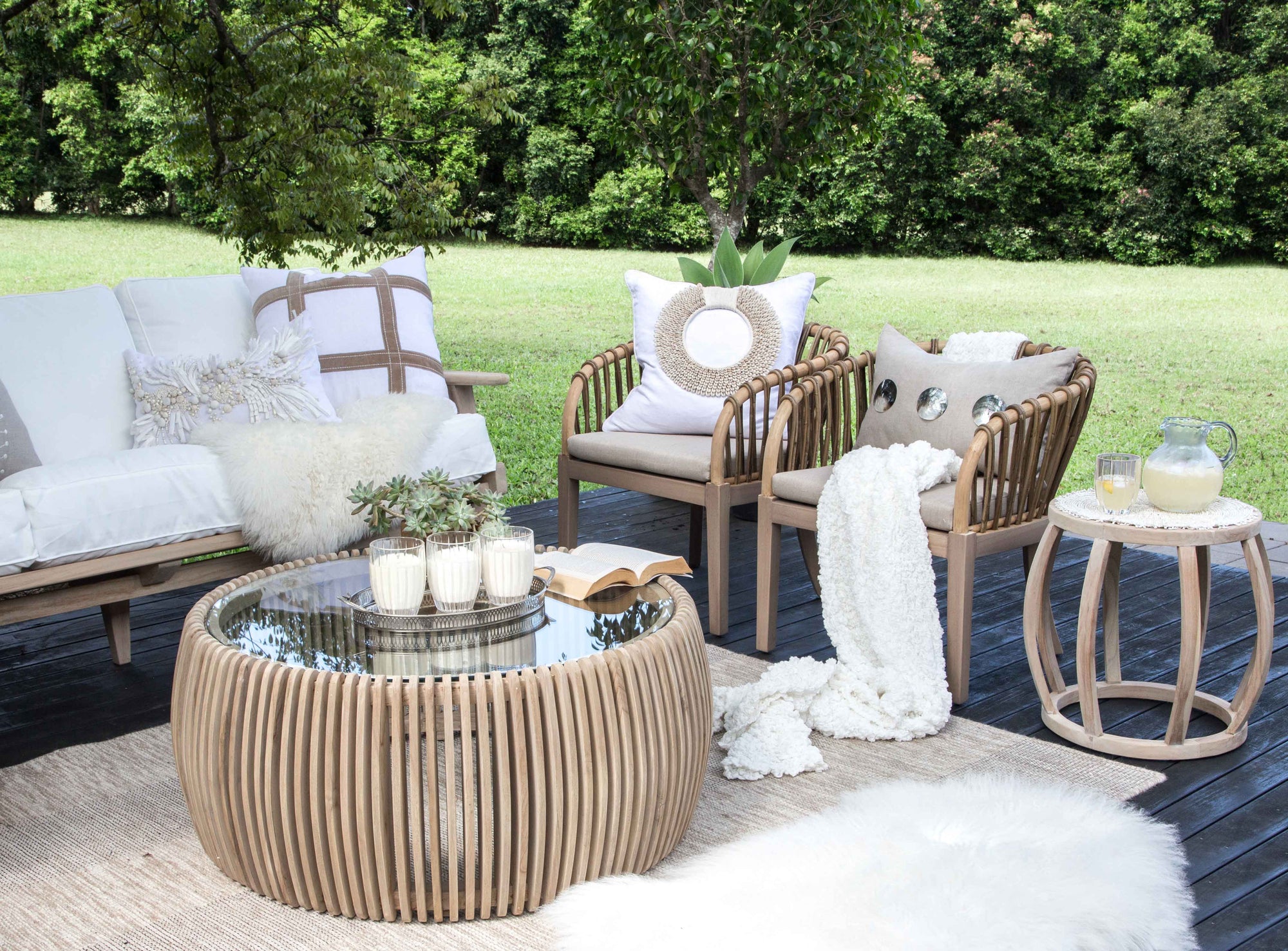 Tranquil outdoor lounge setting featuring the makena Chairs, a teak coffee table, and a cosy white-cushioned sofa styled with soft throws and textured cushions, set against a lush green garden backdrop