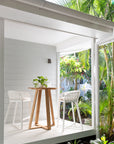 A stylish outdoor bar setting featuring two white Makena bar chairs and a natural timber bar table on a serene verandah. Surrounded by lush greenery, this inviting space embodies the relaxed elegance of slow living