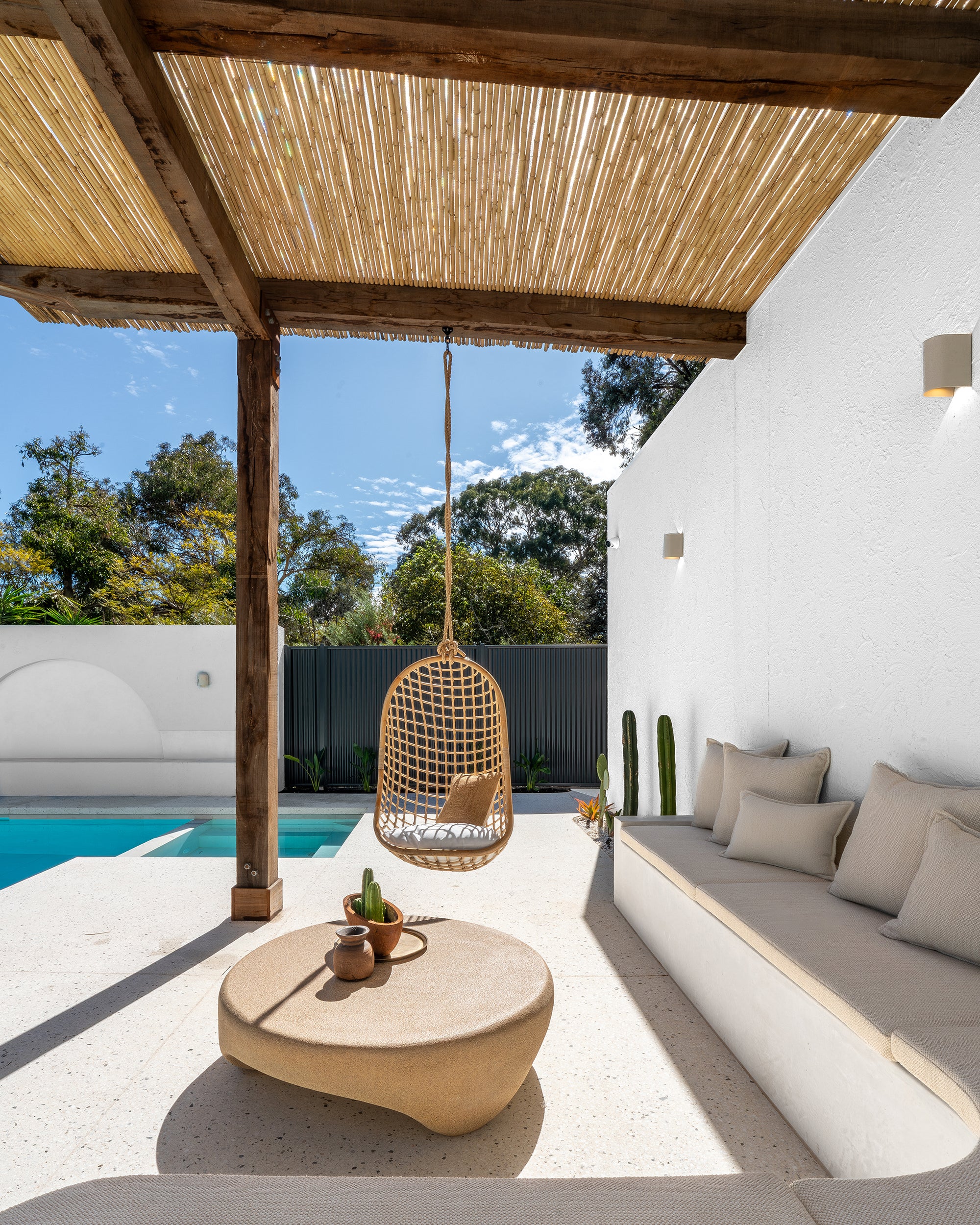 Outdoor poolside lounge with rattan hanging chair, built-in cushioned seating, and sculptural coffee table under a timber pergola.
