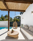 Outdoor poolside lounge with rattan hanging chair, built-in cushioned seating, and sculptural coffee table under a timber pergola.