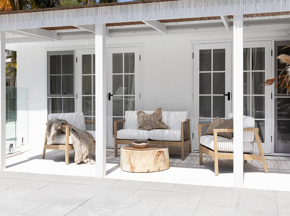 Modern coastal outdoor lounge setting with wooden accent chairs featuring white cushions and sleek spindle detailing, complemented by a round natural wood coffee table and dried floral arrangement