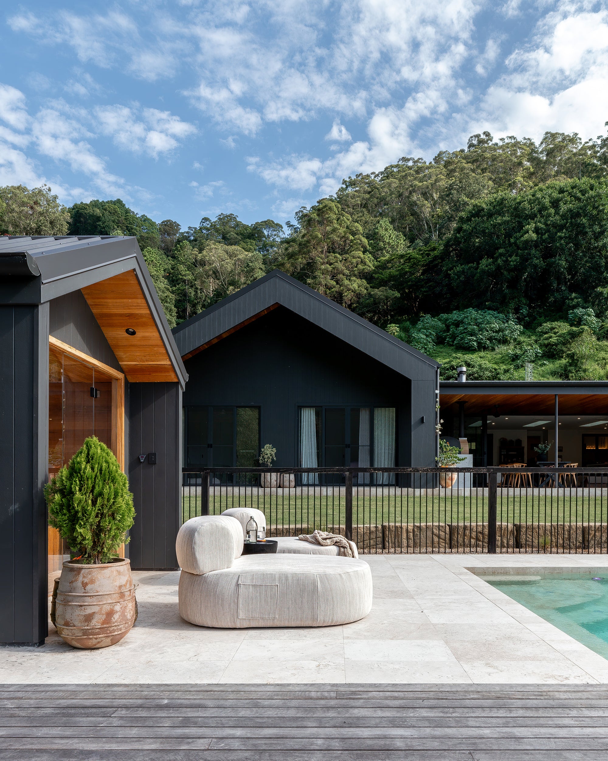 Modern outdoor lounge setting featuring a curved cream upholstered sofa on a tiled deck, set against black timber-clad house with forest backdrop – KULALA Living.