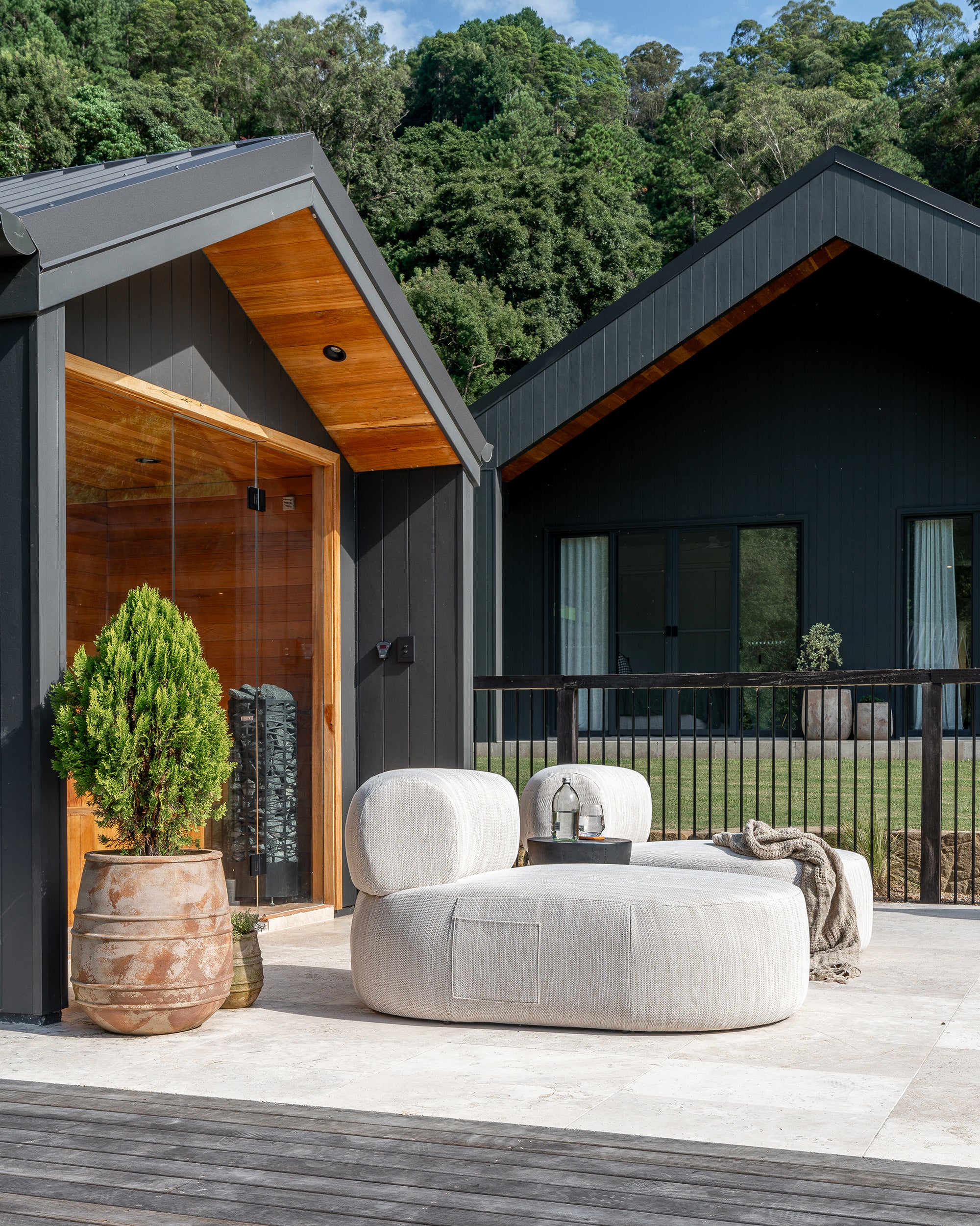 Modern outdoor lounge setting featuring a curved cream upholstered sofa on a tiled deck, set against black timber-clad house with forest backdrop – KULALA Living.