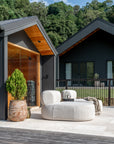 Modern outdoor lounge setting featuring a curved cream upholstered sofa on a tiled deck, set against black timber-clad house with forest backdrop – KULALA Living.
