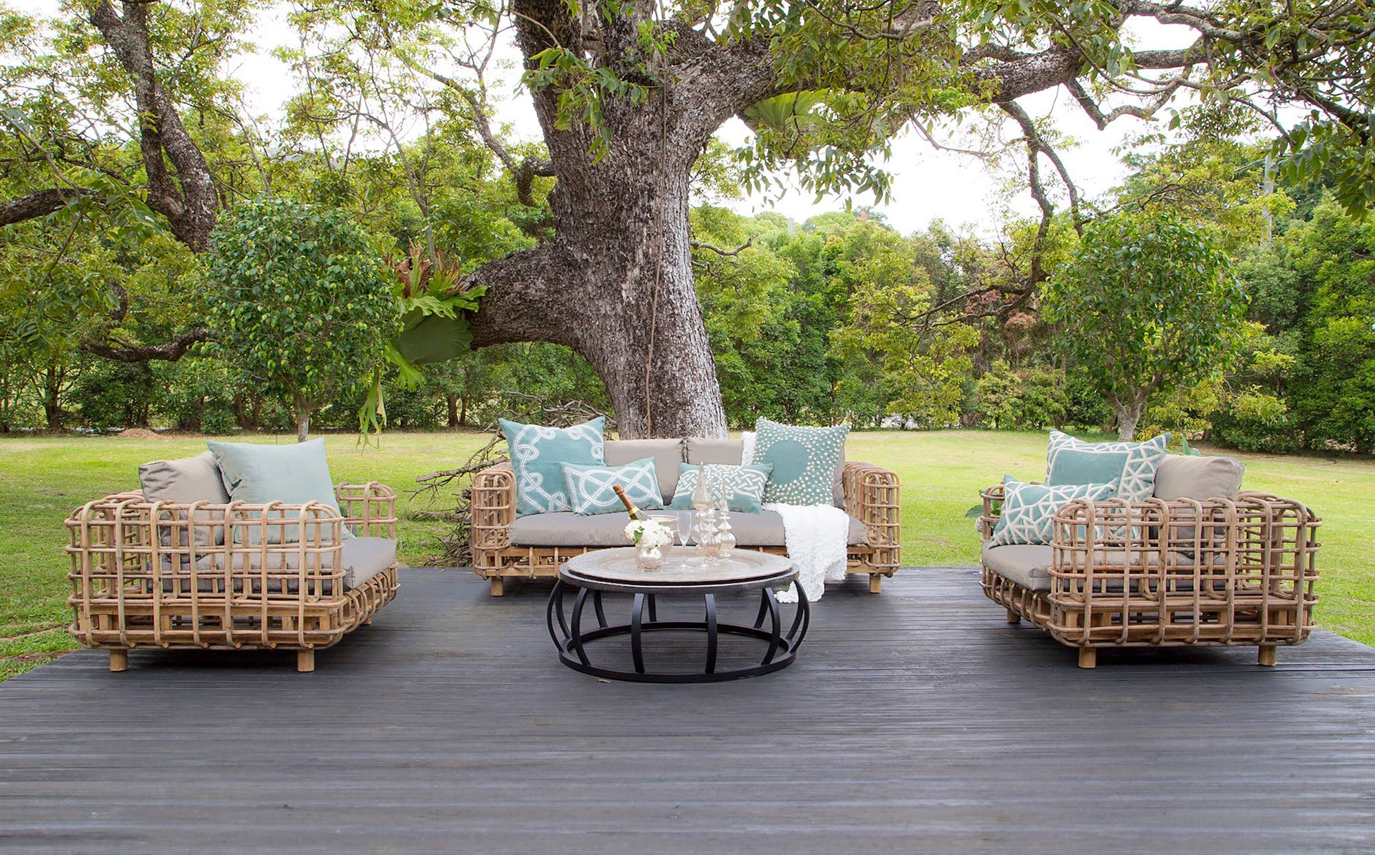 Outdoor lounge setting featuring a handcrafted rattan armchair and sofa with plush grey cushions, styled with soft blue and white accent pillows on a wooden deck under a large tree in a lush garden