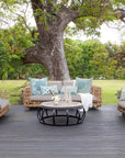 Outdoor lounge setting featuring a handcrafted rattan armchair and sofa with plush grey cushions, styled with soft blue and white accent pillows on a wooden deck under a large tree in a lush garden