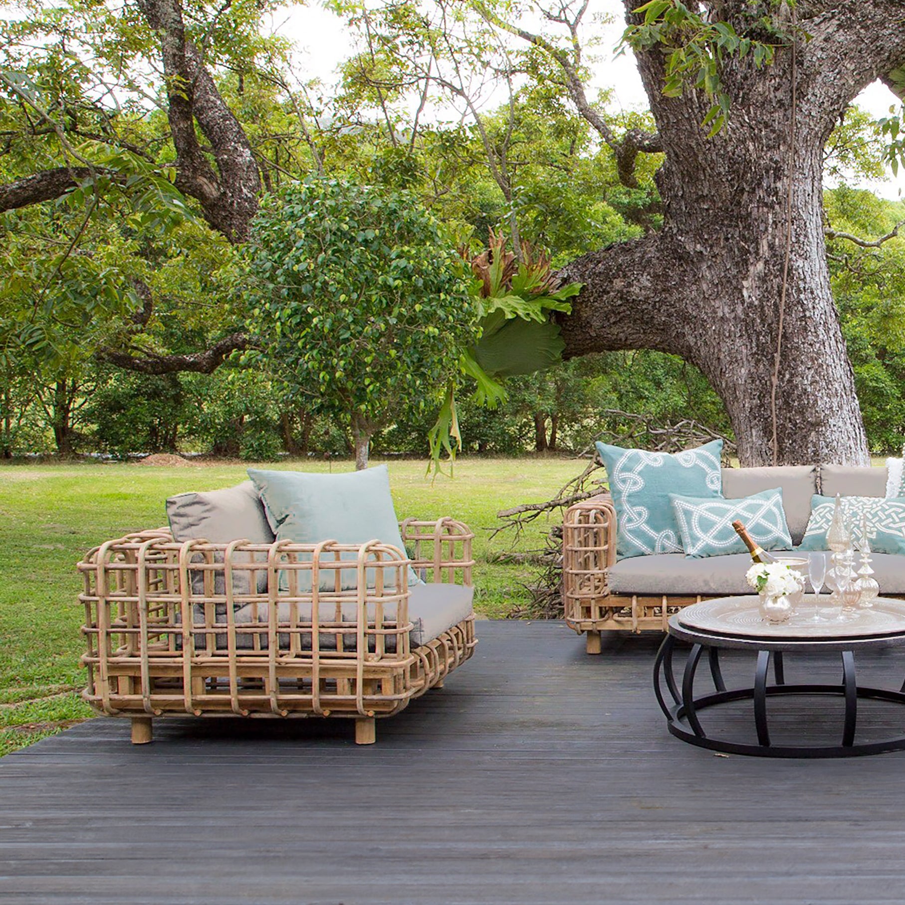 Outdoor lounge setting featuring a handcrafted rattan armchair and sofa with plush grey cushions, styled with soft blue and white accent pillows on a wooden deck under a large tree in a lush garden