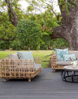 Outdoor lounge setting featuring a handcrafted rattan armchair and sofa with plush grey cushions, styled with soft blue and white accent pillows on a wooden deck under a large tree in a lush garden