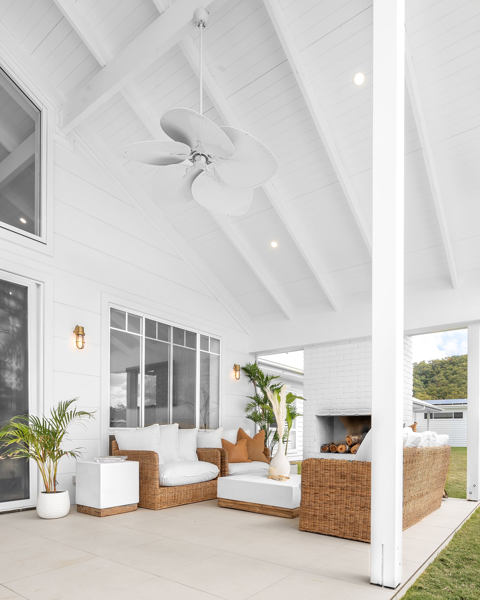 A serene outdoor patio featuring a stylish rattan sofa set with plush white cushions, a modern white coffee table with a wooden base, and a white brick fireplace. Surrounded by lush greenery, this inviting space blends coastal elegance with natural textures, perfect for relaxed outdoor living