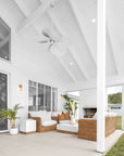 A serene outdoor patio featuring a stylish rattan sofa set with plush white cushions, a modern white coffee table with a wooden base, and a white brick fireplace. Surrounded by lush greenery, this inviting space blends coastal elegance with natural textures, perfect for relaxed outdoor living