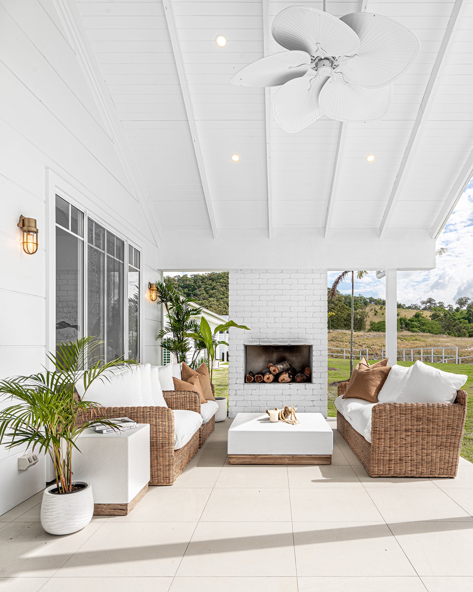 A serene outdoor patio featuring a stylish rattan sofa set with plush white cushions, a modern white coffee table with a wooden base, and a white brick fireplace. Surrounded by lush greenery, this inviting space blends coastal elegance with natural textures, perfect for relaxed outdoor living