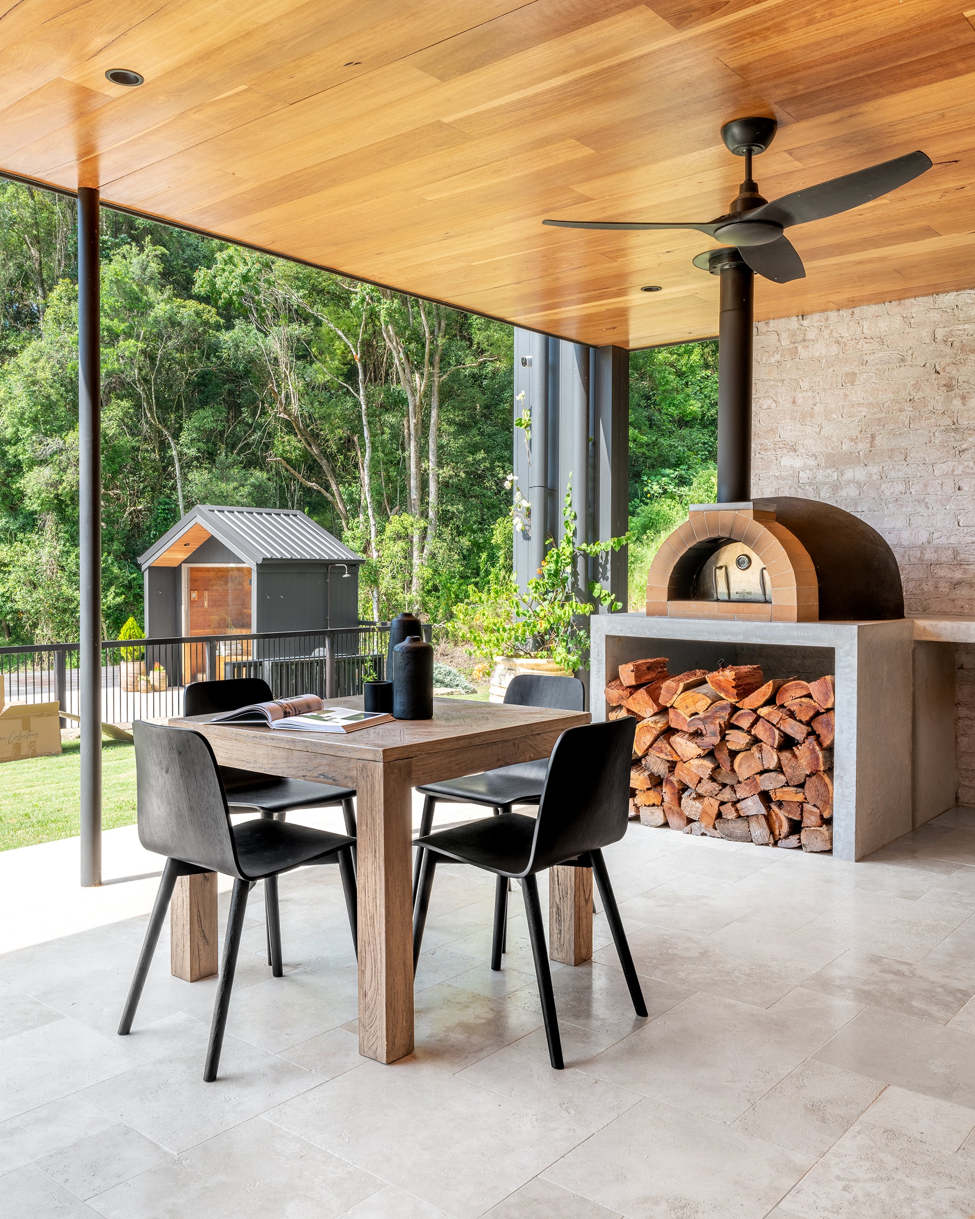 Outdoor dining area with natural timber table, black chairs, and a wood fire oven beneath a timber ceiling, set against lush greenery and modern black timber structures.
