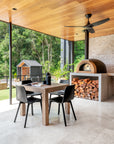 Outdoor dining area with natural timber table, black chairs, and a wood fire oven beneath a timber ceiling, set against lush greenery and modern black timber structures.
