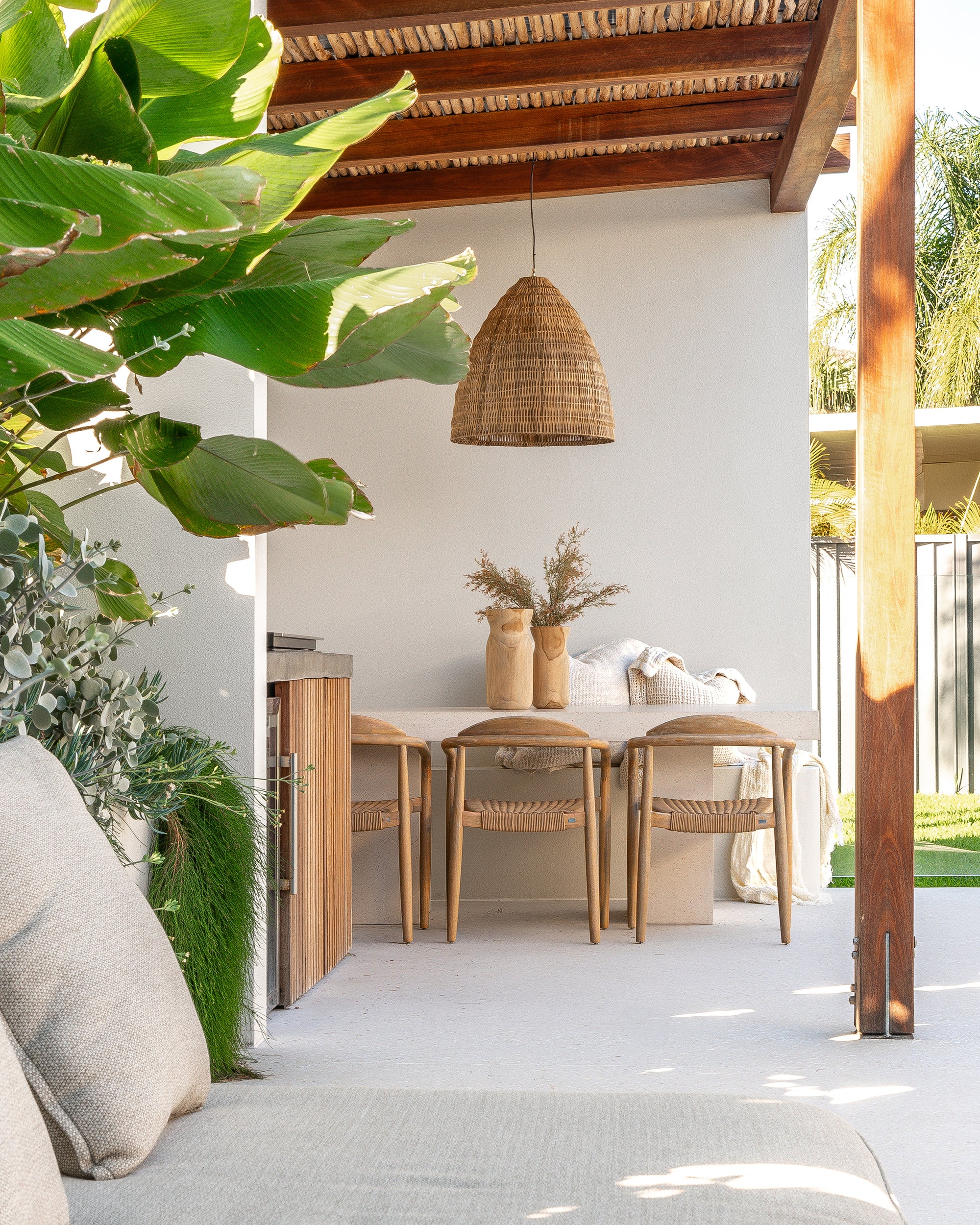Outdoor dining setting featuring a teak chairs and travertine-look dining table beside a pool under a woven pendant light