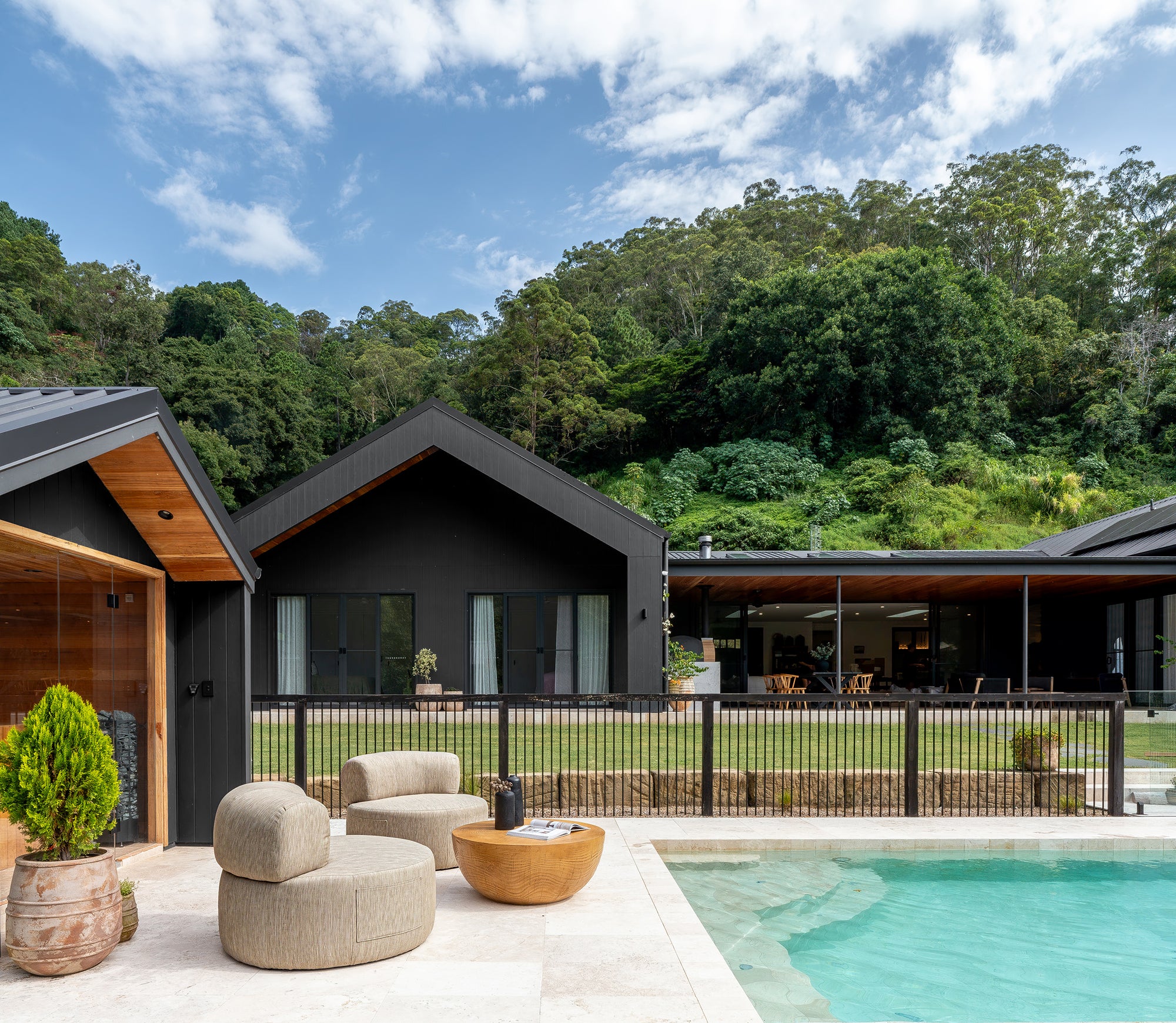 Outdoor setting featuring rounded occasional chairs and sculptural magnesium oxide coffee table by a poolside, with a modern black-clad pavilion home set against a lush hinterland backdrop