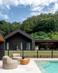 Outdoor setting featuring rounded occasional chairs and sculptural magnesium oxide coffee table by a poolside, with a modern black-clad pavilion home set against a lush hinterland backdrop