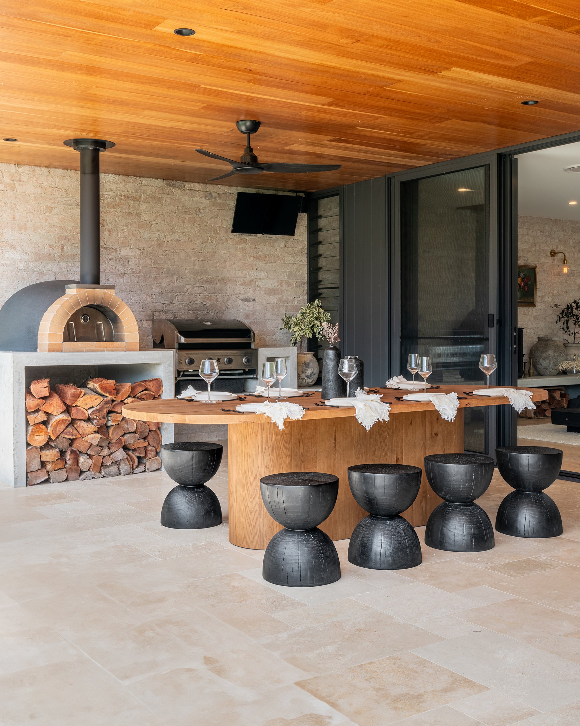 Outdoor dining area featuring black hourglass stools, solid timber dining table, and a built-in woodfire pizza oven beneath a warm timber ceiling.