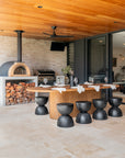 Outdoor dining area featuring black hourglass stools, solid timber dining table, and a built-in woodfire pizza oven beneath a warm timber ceiling.