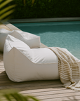 Playa Chair in white outdoor fabric styled beside a pool on timber decking with a striped towel draped over the seat in soft natural light.