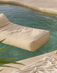 Playa Lounger in sand colour floating in an organic-shaped pool with soft rippling water, styled with a fringed towel and sandals beside the poolside.