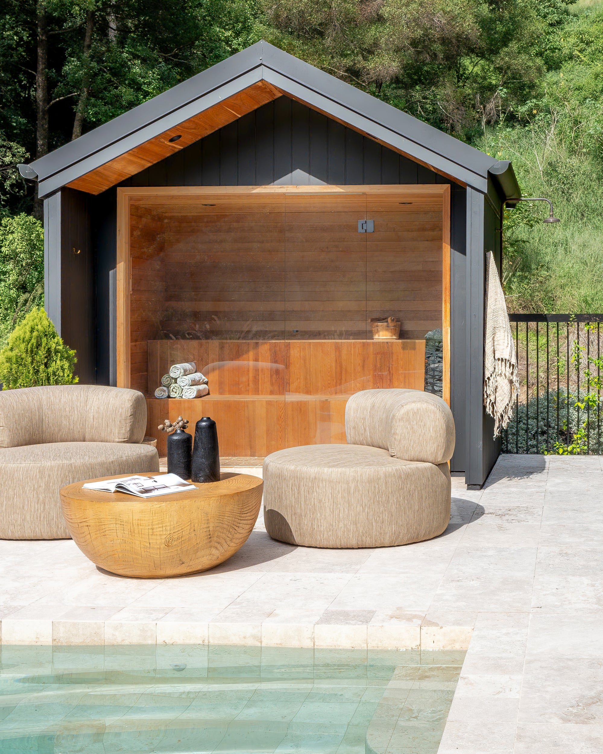 Poolside lounge area featuring two contemporary armless outdoor chairs in textured neutral fabric and a solid round magnesium oxide coffee table, set in front of a timber-lined sauna with glass doors.
