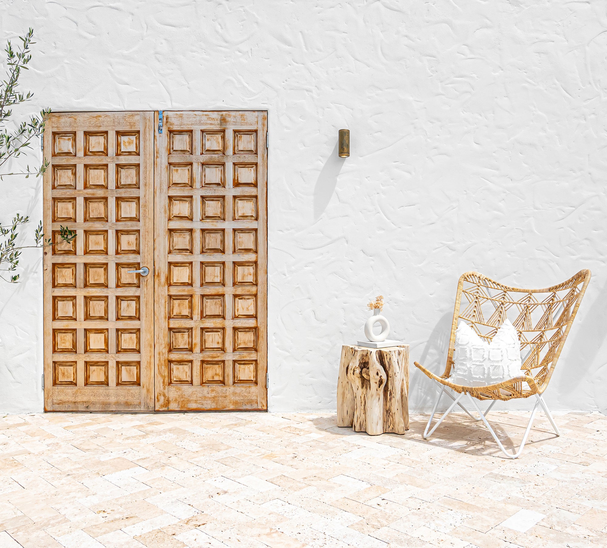A minimalist outdoor setting featuring a rattan butterfly chair with a white metal frame, styled with a textured cushion. Paired with a natural tree stump side table holding a ceramic vase, set against a rustic timber door and white textured wall for a Mediterranean-inspired aesthetic