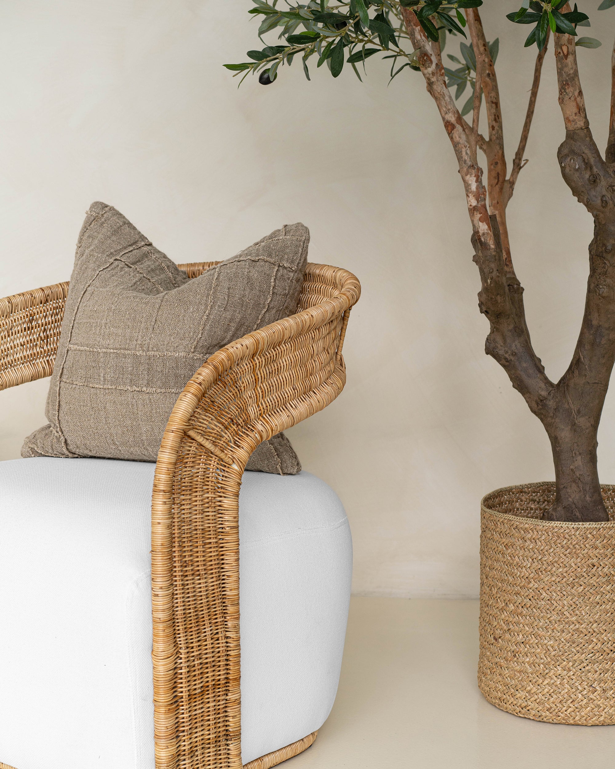 Natural rattan armchair with a white cushion, styled with a textured linen throw pillow in earthy tones, next to a woven basket planter with an olive tree