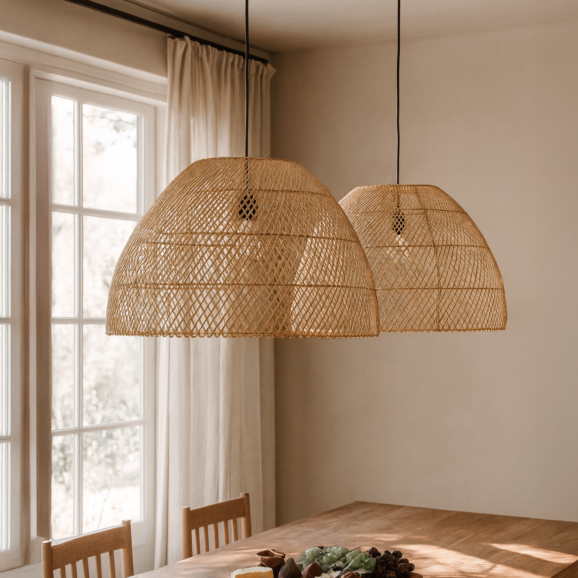 Pair of woven rattan pendant lights hanging above a wooden dining table with a cheese and fruit platter, styled in a sunlit dining room.