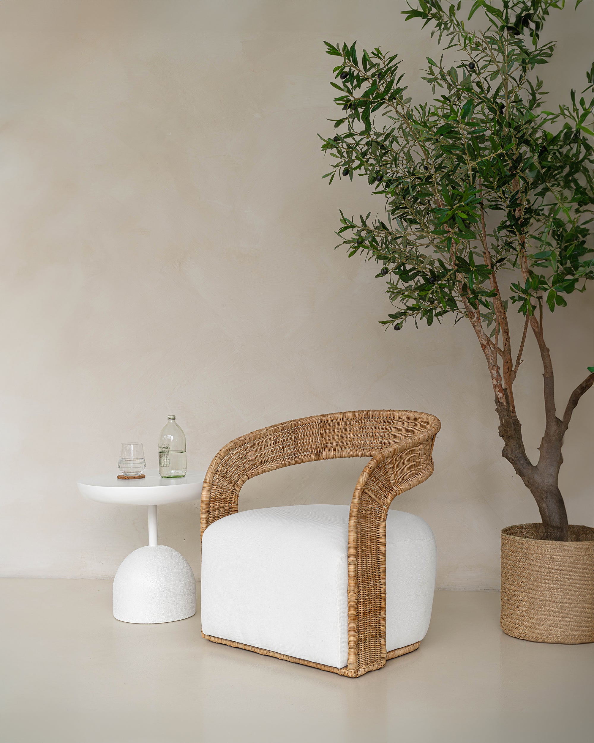 A close-up of a rattan armchair with a white upholstered seat, styled with a textured linen cushion. Paired with a woven basket planter and an olive tree, this setting exudes natural elegance and relaxed sophistication