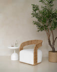 A close-up of a rattan armchair with a white upholstered seat, styled with a textured linen cushion. Paired with a woven basket planter and an olive tree, this setting exudes natural elegance and relaxed sophistication