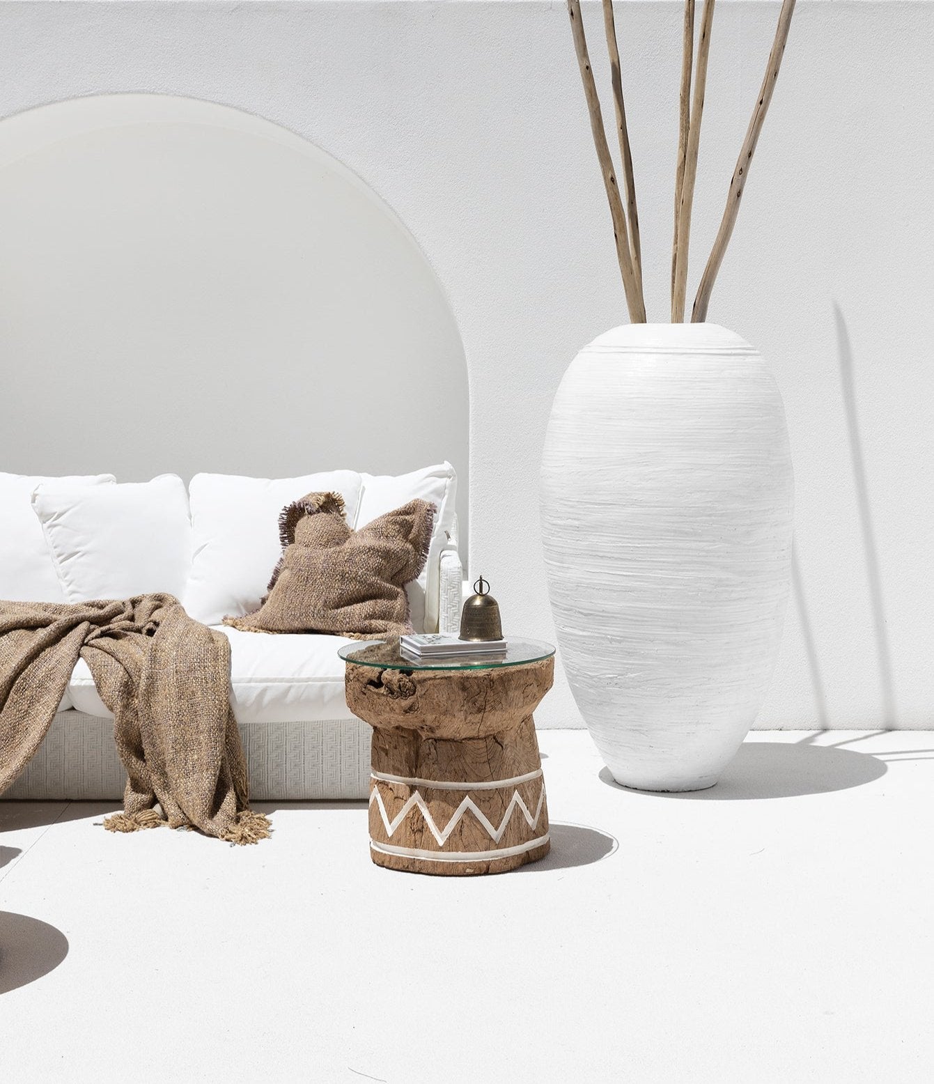 A reclaimed teak side table with a glass top, featuring white tribal-inspired detailing, styled in an outdoor setting. Paired with a white sculptural vase and a woven daybed adorned with textured cushions and throws, this serene space embodies relaxed, coastal-inspired living