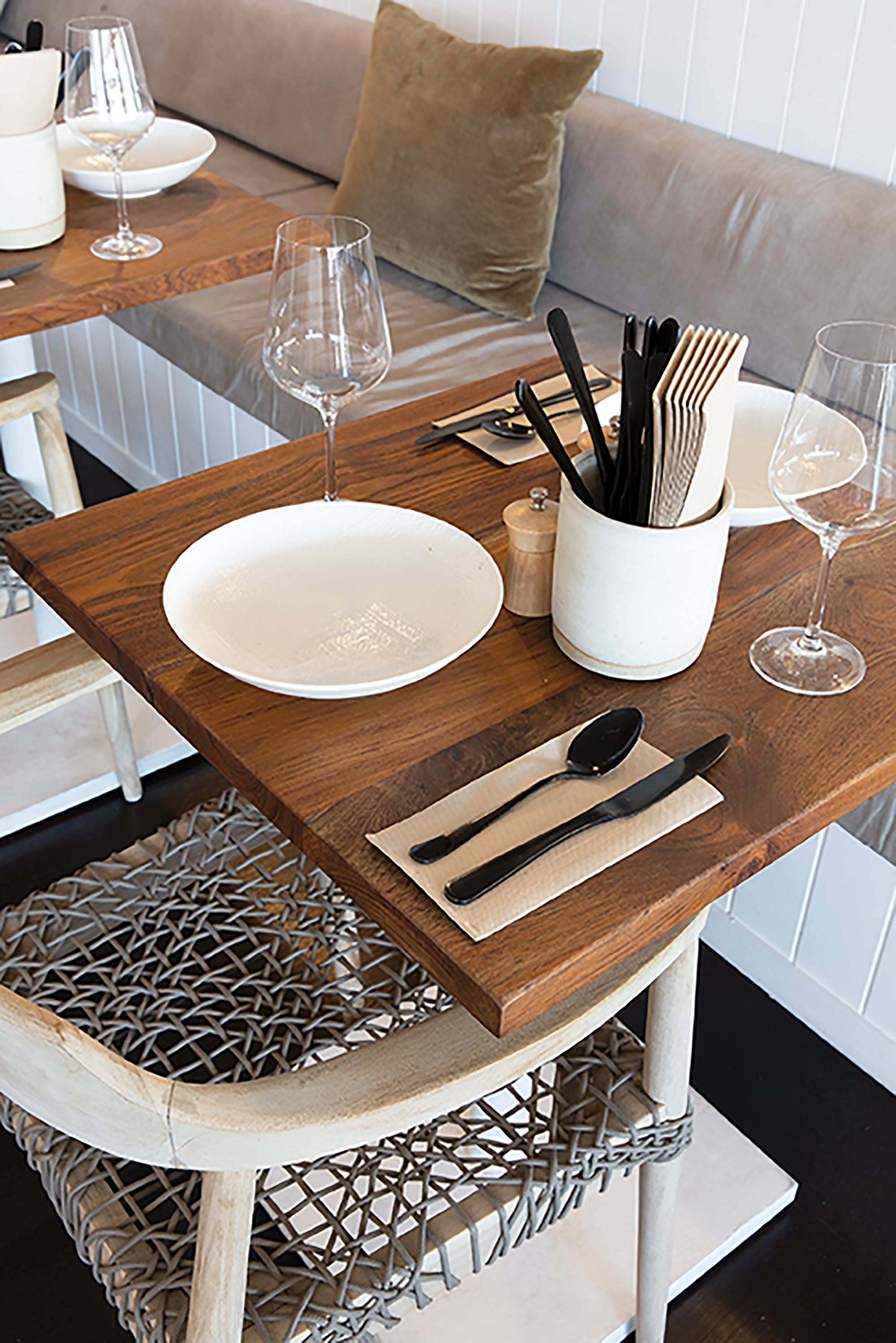 Close-up of a restaurant dining setup with a wooden table, woven rope dining chair, white ceramic plate, black cutlery, wine glasses, and a neutral cushion on a bench seat.