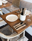 Close-up of a restaurant dining setup with a wooden table, woven rope dining chair, white ceramic plate, black cutlery, wine glasses, and a neutral cushion on a bench seat.