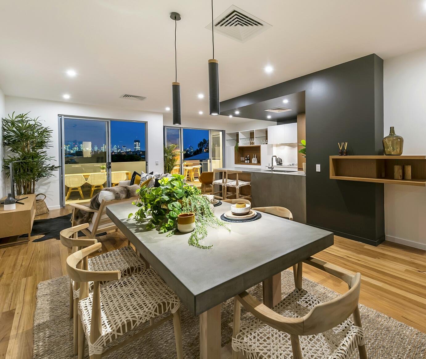 Contemporary dining room with a concrete dining table and wooden chairs featuring woven seats, overlooking a city skyline at night through large glass doors.