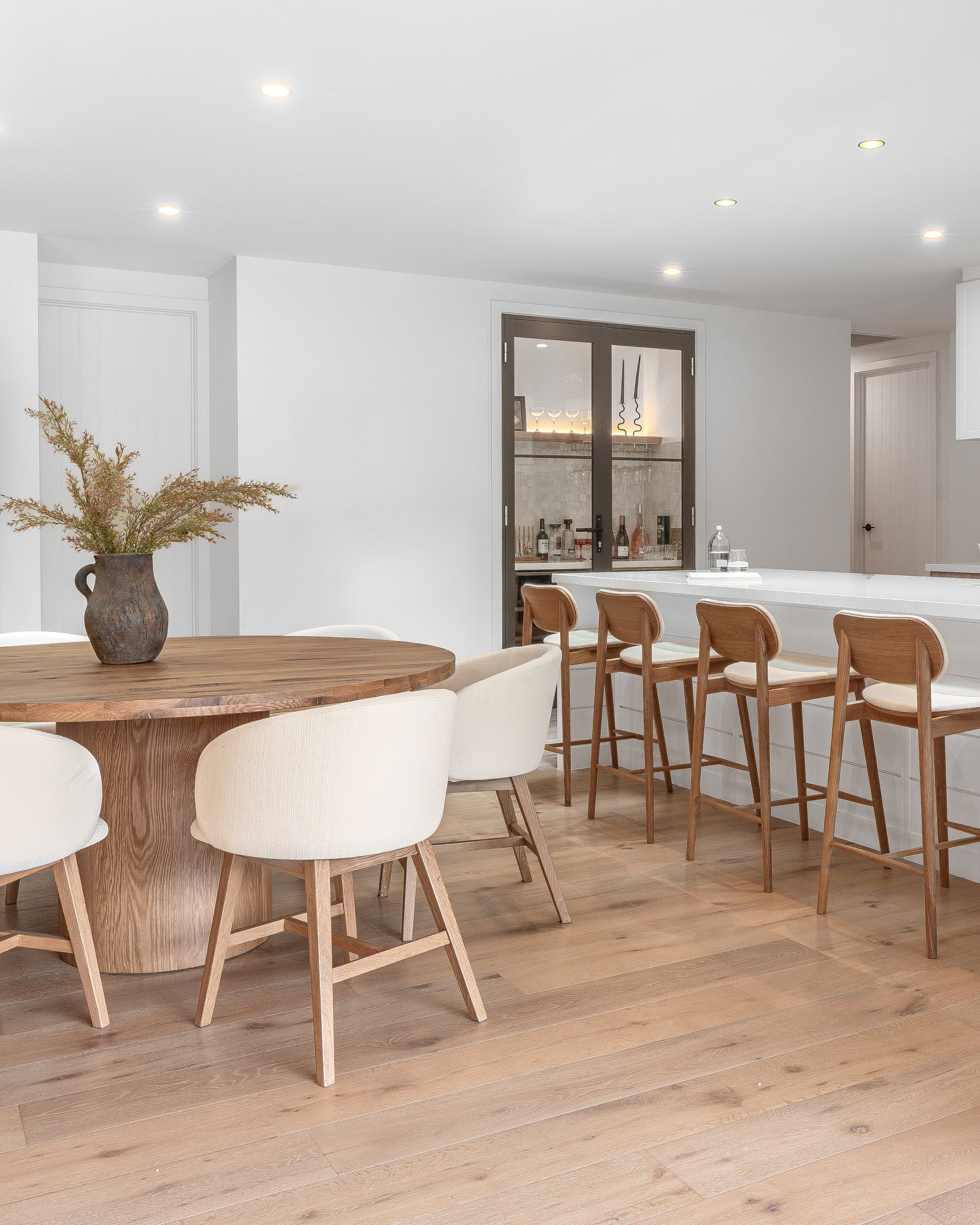 Contemporary dining space featuring a round oak dining table with upholstered chairs and matching oak bar stools at a white kitchen island, styled with neutral tones and soft textures.