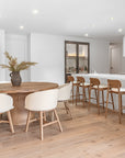 Contemporary dining space featuring a round oak dining table with upholstered chairs and matching oak bar stools at a white kitchen island, styled with neutral tones and soft textures.